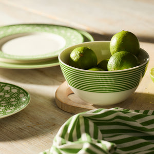 A Newport Stripe Verde Cereal Bowl by Caskata, filled with limes, rests on a wooden surface beside stacked green and white plates, accented by a Newport Stripe cloth in the foreground.