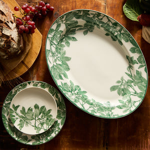 The Arbor Green Oval Rimmed Platter by Caskata Artisanal Home graces the wooden table with its green leaf patterns, embodying both classic and contemporary elegance. Accompanied by a loaf of bread, grapes, and figs, this setting defines high style in dining.