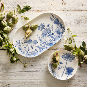 The Summer Blues Large Coupe Oval Platter by Caskata, with its blue floral patterns, is elegantly placed on a rustic wooden table, surrounded by artichokes and leaves.