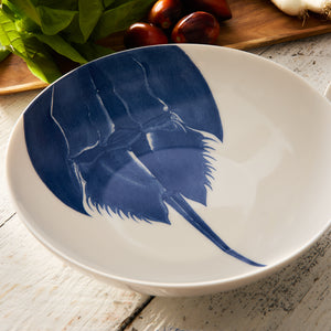 The Horseshoe Crab Wide Serving Bowl from Caskata Artisanal Home features a subtle blue horseshoe crab design on its base, crafted from high-fired porcelain. It is beautifully displayed on a wooden surface next to leafy greens and tomatoes.