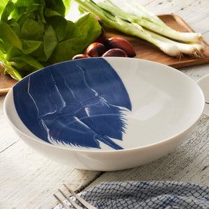 A Horseshoe Crab Wide Serving Bowl by Caskata Artisanal Home, featuring a blue horseshoe crab design, sits next to a salad, green onions, chestnuts, and a fork on a wooden surface.