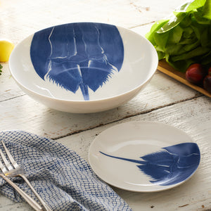 The Caskata Artisanal Home Horseshoe Crab Wide Serving Bowls, skillfully crafted from high-fired porcelain, showcase intricate blue horseshoe crab designs and sit gracefully on a wooden table. Nearby lie fresh lettuce, tomatoes, a lemon, a checkered napkin, and a fork.