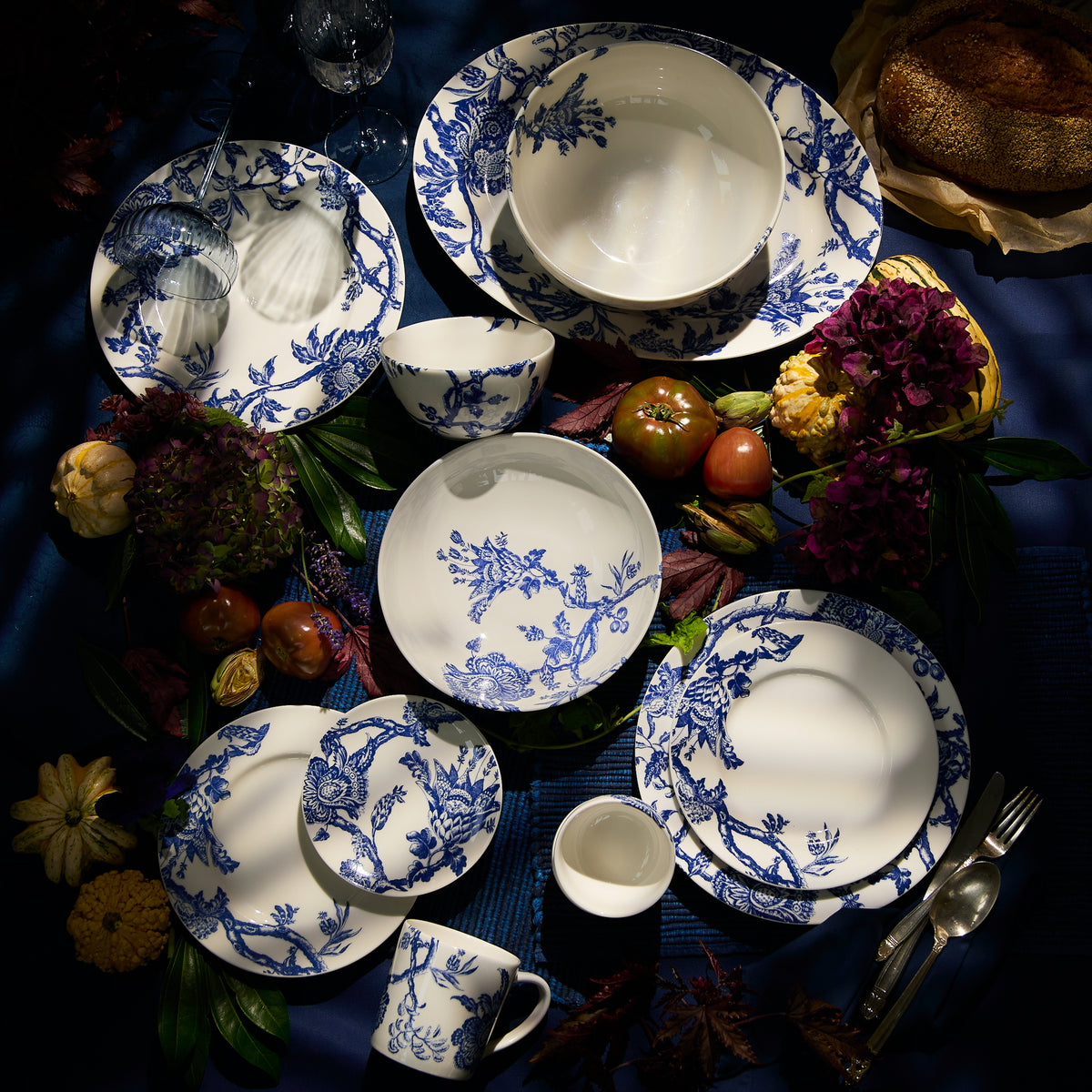 A table set with the Arcadia Oval Rimmed Platter from Caskata Artisanal Home, showcasing premium porcelain featuring a blue and white floral pattern, surrounded by assorted flowers, fruits, and bread under soft lighting.