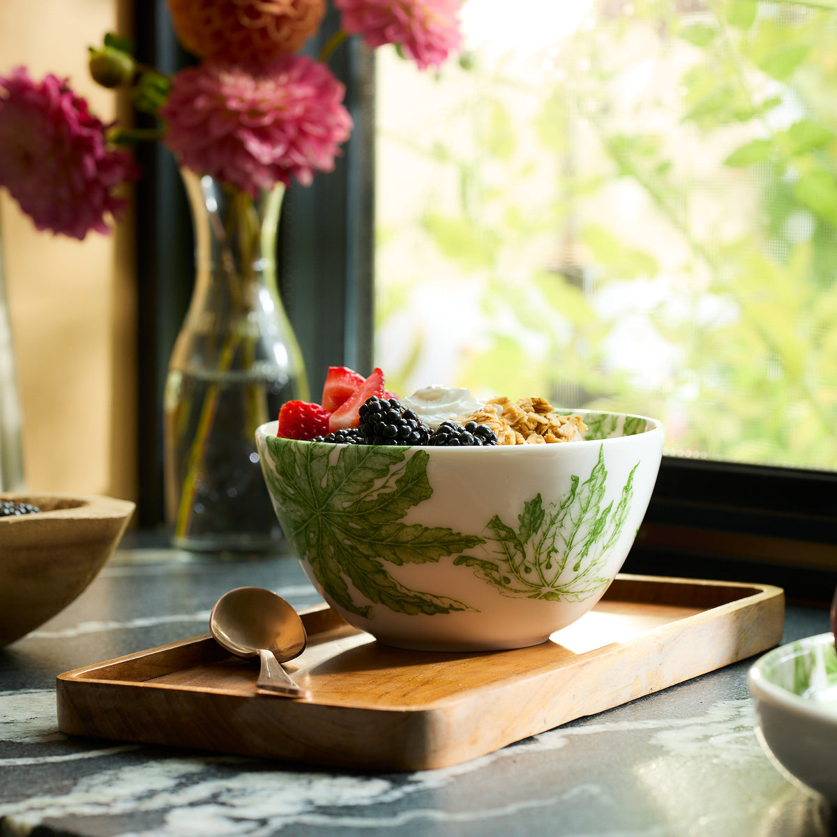 A Freya Cereal Bowl by Caskata, featuring a green leaf design, sits on a wooden tray. It is filled with yogurt, granola, and berries. A spoon rests beside it while pink flowers beautify a vase in the background.