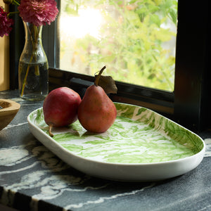 Two pears are elegantly placed on the Freya Large Coupe Oval Platter by Caskata, resting on a marble countertop near a window with green florals beside them. This charming arrangement is an entertaining must-have that adds elegance to any gathering.