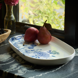 Two red pears rest on the Summer Blues Large Coupe Oval Platter by Caskata, crafted from high-fired porcelain and showcasing a beautiful blue floral design. The arrangement sits elegantly on a marble countertop near a window, blending perfectly with the summer blues of the surrounding decor.