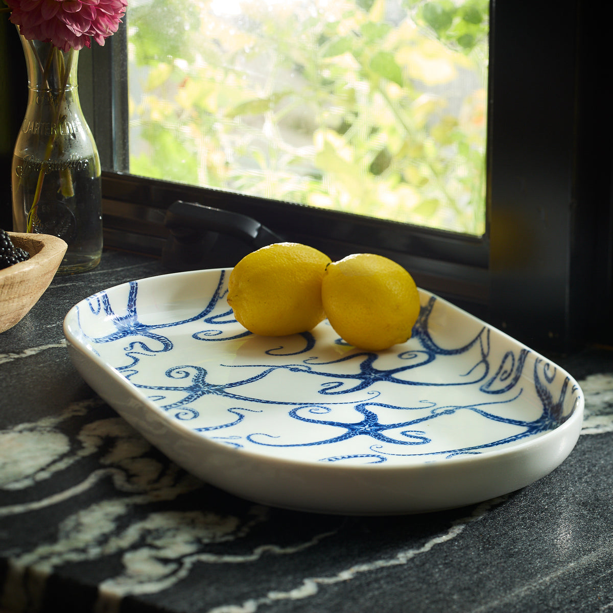 The Starfish Large Coupe Oval Platter by Caskata, adorned with blue swirl patterns, holds two lemons on a black marble countertop near a window framed by lush greenery, evoking the essence of sea stars.