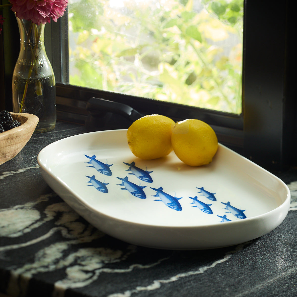 The School of Fish Large Coupe Oval Platter by Caskata, featuring blue fish designs, elegantly holds two lemons on a textured countertop. Nearby, a window showcases a vase of flowers and a basket of blackberries, making it an entertaining must-have for any dinnerware collection.