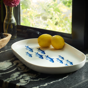 The School of Fish Large Coupe Oval Platter by Caskata, featuring blue fish designs, elegantly holds two lemons on a textured countertop. Nearby, a window showcases a vase of flowers and a basket of blackberries, making it an entertaining must-have for any dinnerware collection.