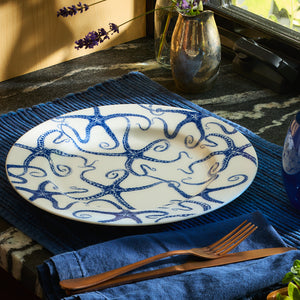 Limited-edition Starfish Rimmed Dinner Plate from Caskata Artisanal Home, featuring a blue octopus tentacle pattern, stylishly arranged on a blue placemat with a fork and knife.