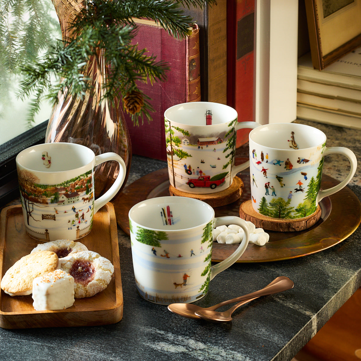 The table features a set of four Caskata X Felix Doolittle Winter Wonderland Mugs, each decorated with charming winter scenes. A copper plate nearby holds cookies, sugar cubes, and two spoons. In the background, a plant and books enhance the cozy New England atmosphere.