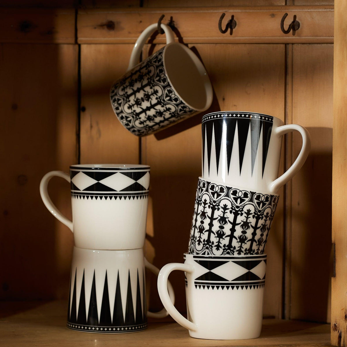 A collection of Casablanca Mugs from Caskata Artisanal Home, featuring black geometric patterns, is arranged on a wooden shelf.