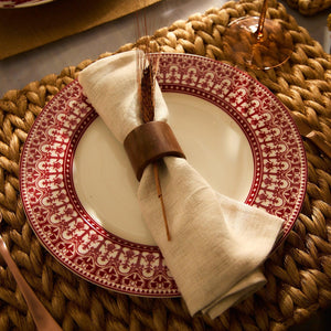 A Casablanca Crimson Rimmed Dinner Plate from Caskata Artisanal Home, accompanied by a beige cloth napkin secured with a wooden ring, set on a woven placemat.