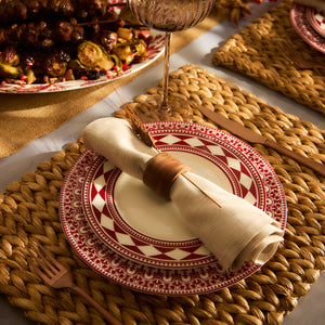 A dining table arrangement showcases a Caskata Artisanal Home Fez Crimson Rimmed Salad Plate with a Moroccan pattern, accompanied by a rolled napkin in a holder and a fork on a woven placemat. In the background, skewered food and a glass capture the enduring allure of Fez geometrics.