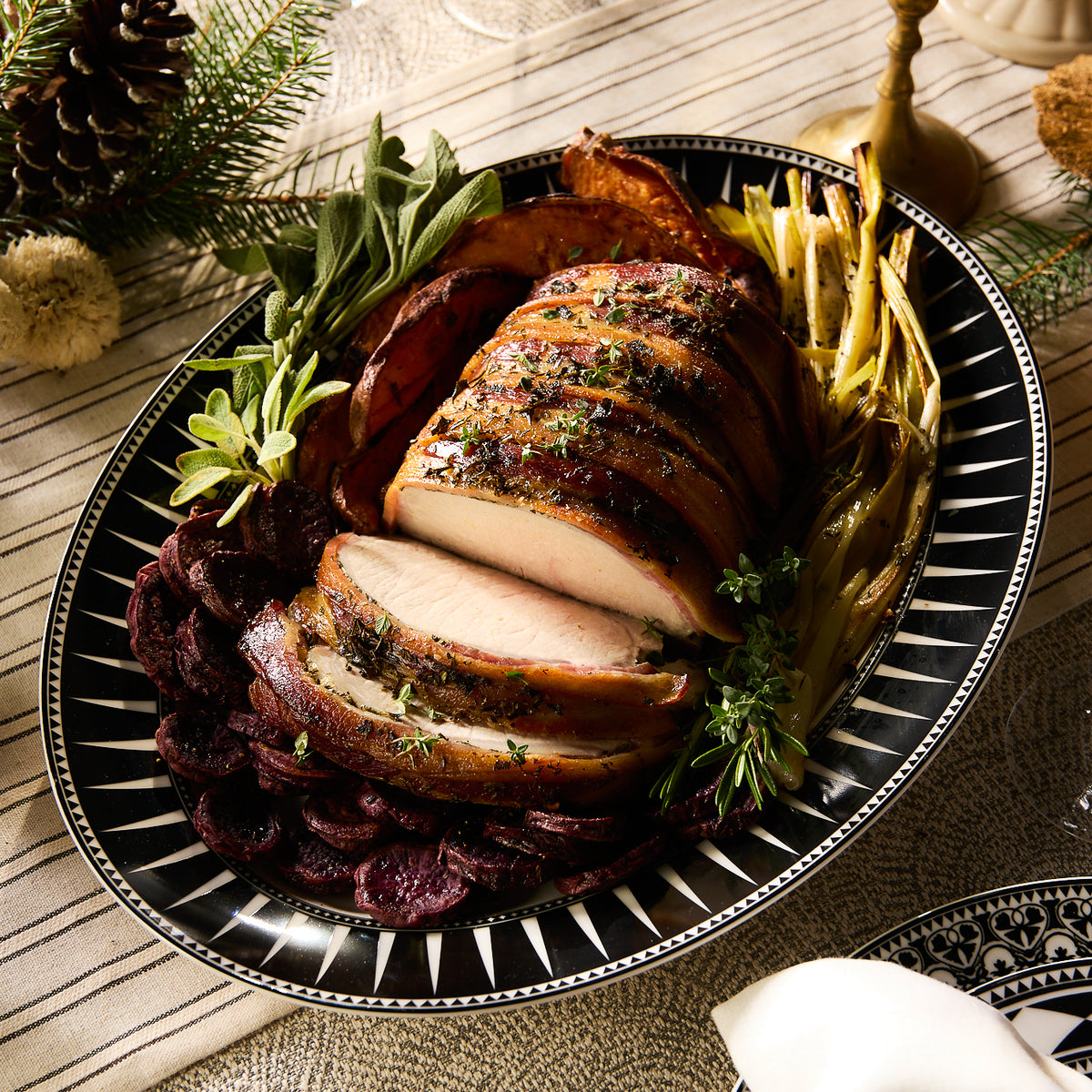 A roasted pork loin with herbs is elegantly sliced and served on a decorative Marrakech Oval Rimmed Platter by Caskata Artisanal Home, accompanied by roasted beets and vegetables.