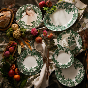 A table setting featuring Caskata's Arbor Green Rimmed Dinner Plate adorned with a green and white floral pattern, surrounded by an assortment of fruits, vegetables, bread, and flowers on a patterned tablecloth highlights intricate botanical details.