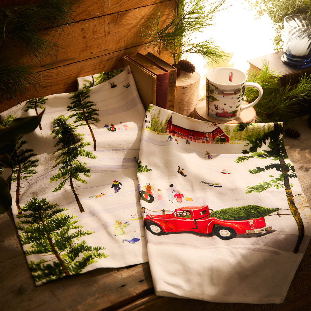 A cozy scene comes to life with the Caskata Winter Wonderland Kitchen Towels, showcasing a charming winter landscape of sledding children alongside a red truck, a mug, books, and pine branches. Warm lighting adds to the rustic ambiance.