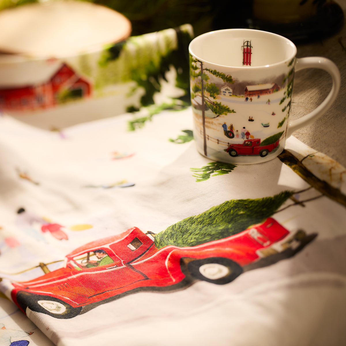 A Caskata festive mug featuring a winter scene is placed on a tablecloth decorated with a red truck carrying a Christmas tree, beautifully enhancing the Caskata X Felix Doolittle Winter Wonderland Kitchen Towels, Set of 2.