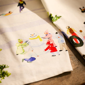 A close-up of the Caskata X Felix Doolittle Winter Wonderland Kitchen Towels, set of 2, showcases charming illustrations of children building a snowman and sledding against a backdrop of trees and snow—making it a perfect holiday gift.