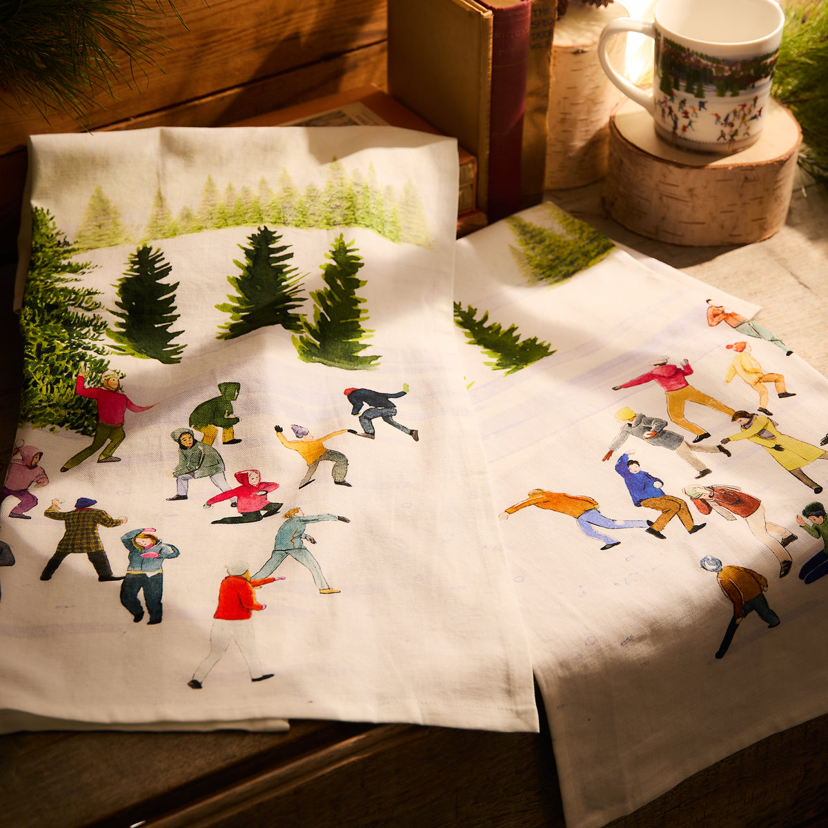 The Caskata X Felix Doolittle Snowball Fun Kitchen Towels, in a set of 2, showcase vibrant winter scenes of people ice skating and trees. They are displayed on a wooden surface, alongside a cozy mug and a stack of books.