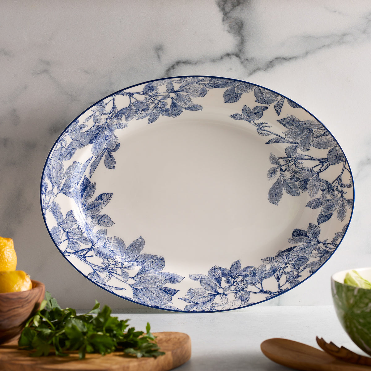 The elegant Arbor tableau features the Arbor Oval Rimmed Platter by Caskata Artisanal Home, a high-style porcelain piece with blue floral patterns, resting on a marble surface amidst fresh herbs, lemons, and a wooden bowl.