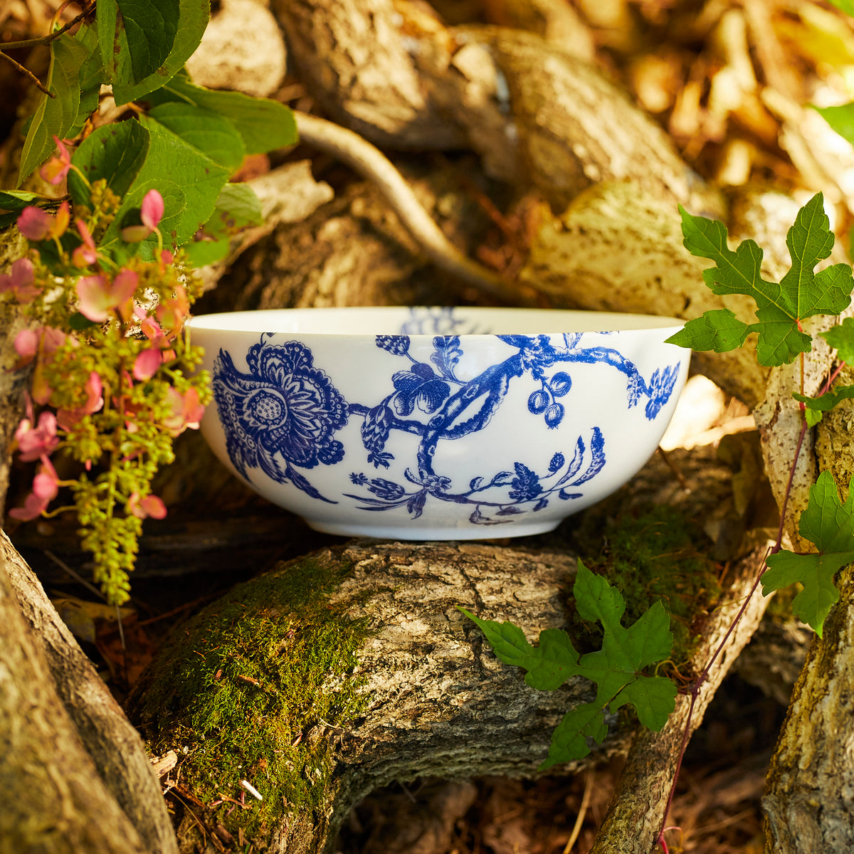 An Arcadia Vegetable Serving Bowl by Caskata Artisanal Home with blue floral patterns rests on a twisted tree branch, surrounded by green leaves and pink flowers. This exquisite piece is inspired by designs from the Williamsburg Foundation.
