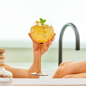 A hand gently holds a Quinn Amber Red Wine Glass by Caskata Artisanal Home, showcasing a yellow drink with mint garnish, elegantly positioned near the bathtub faucet.