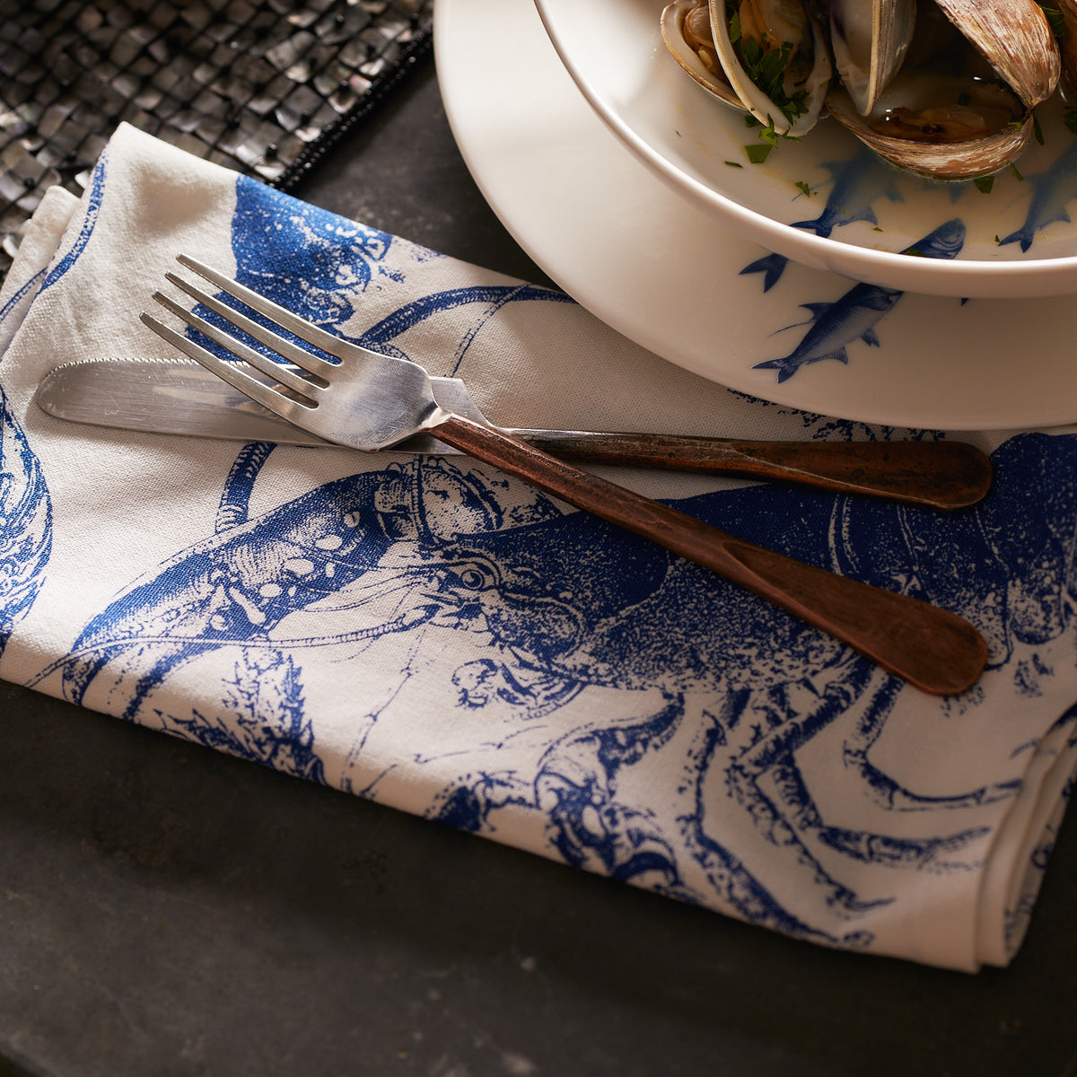 A fork and knife rest on a patterned napkin alongside a bowl of clams, perfectly suited for any Caskata Lobster Boil Bundle set.