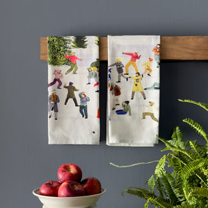 The set of two Caskata X Felix Doolittle Snowball Fun Kitchen Towels, featuring vibrant illustrations of joyful figures, are elegantly draped on a wooden bar. A bowl filled with red apples and a lush green fern sit in the foreground, infusing this cozy kitchen scene with a natural charm.