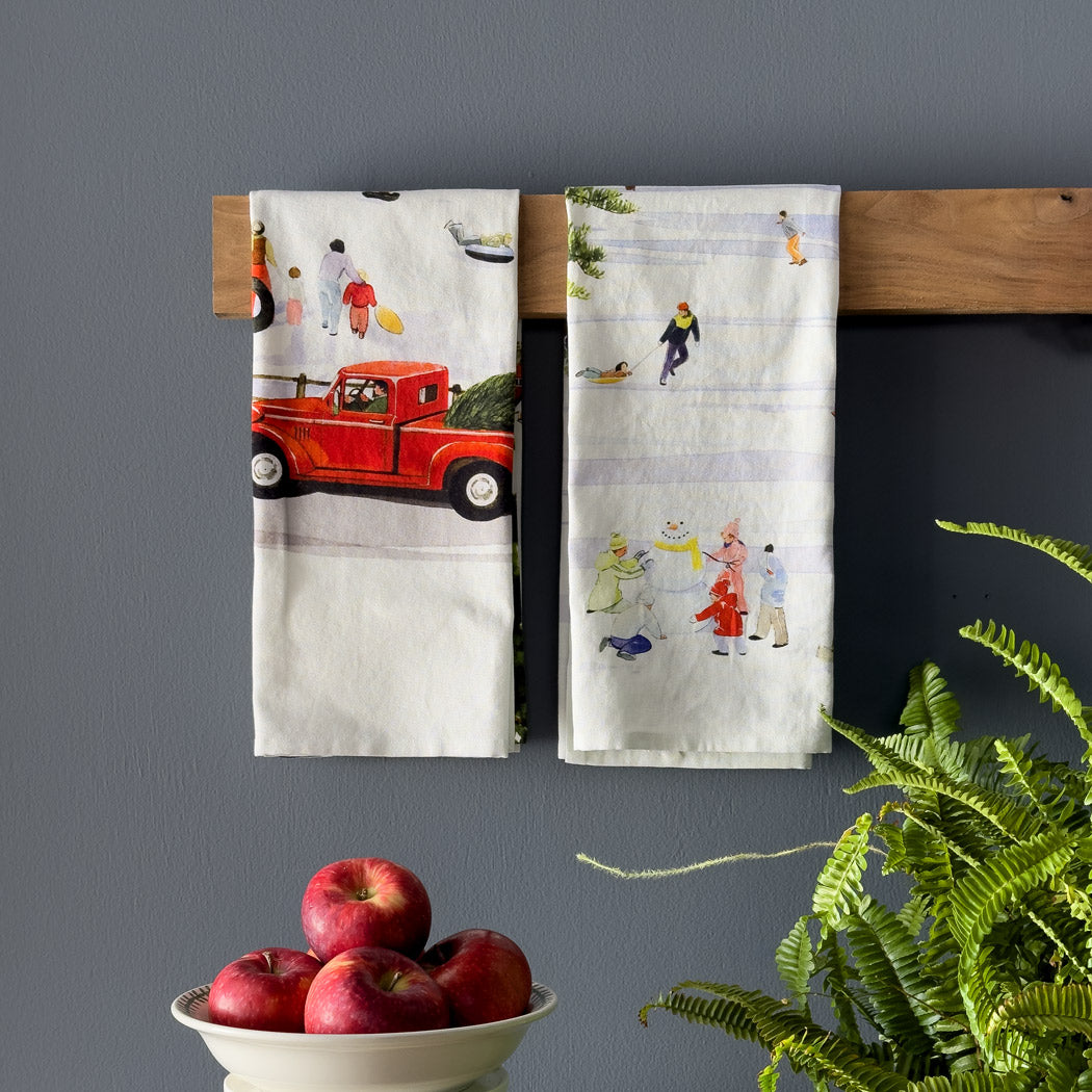 Two Caskata X Felix Doolittle Winter Wonderland Kitchen Towels, featuring vintage-themed prints, hang on a wooden rack. Below them sits a bowl filled with red apples and accompanied by a green plant, creating a charming and inviting display.