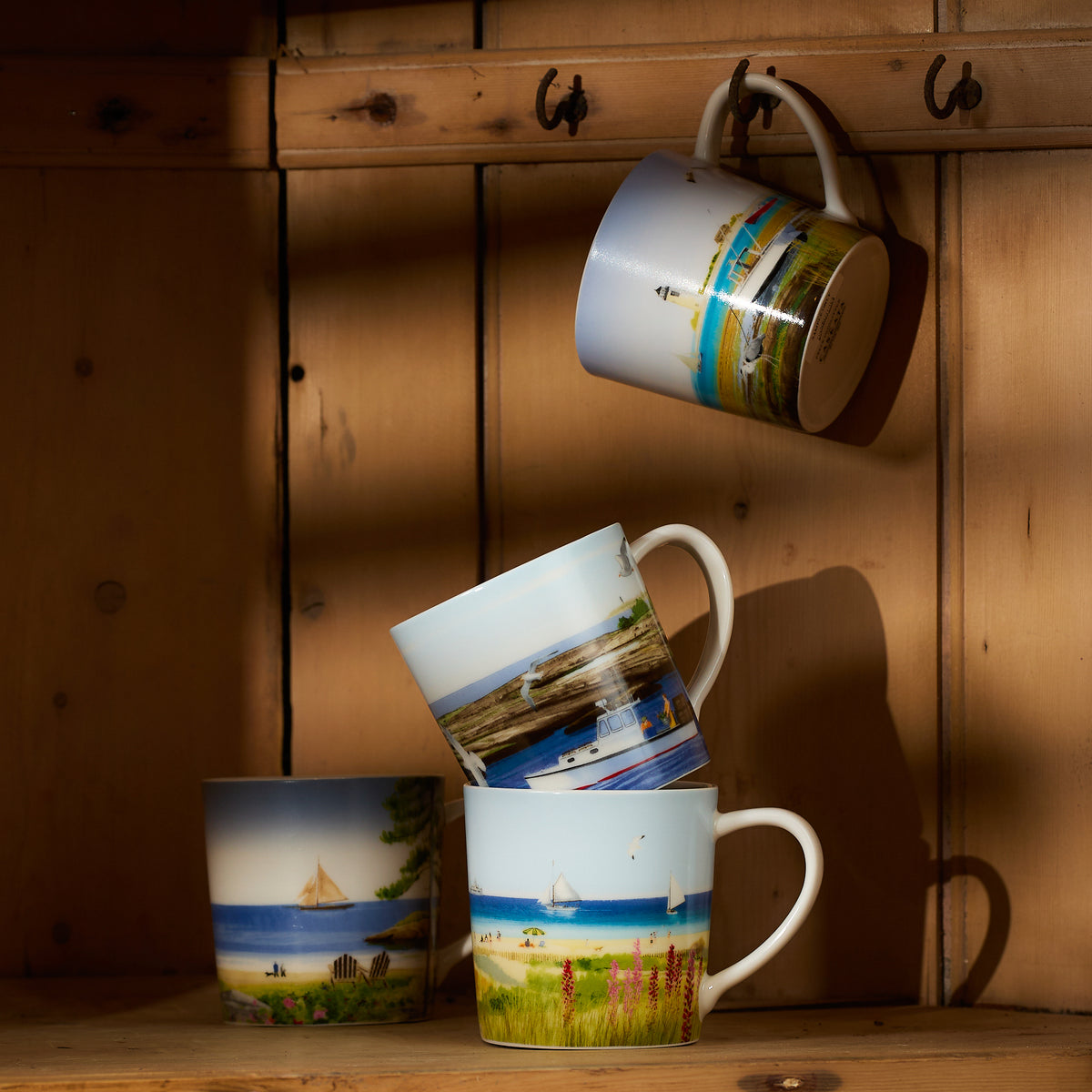 The Caskata X Felix Doolittle By the Sea Mugs, a set of four, are beautifully stacked and hanging on wooden hooks in a sunlit wooden shelf.