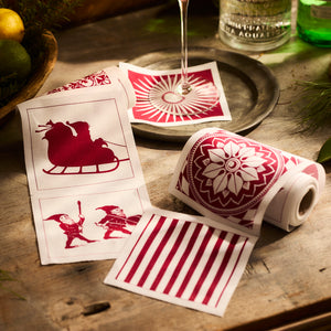 Holiday-themed cotton coaster napkins showcasing festive patterns, including deep red designs such as sleds, elves, floral motifs, and stripes. Placed on a wooden surface with lemons and a drink nearby, these coasters set the perfect scene for your Holiday. Fun Cocktail Napkin Rolls from Caskata are sold in a set of 2.