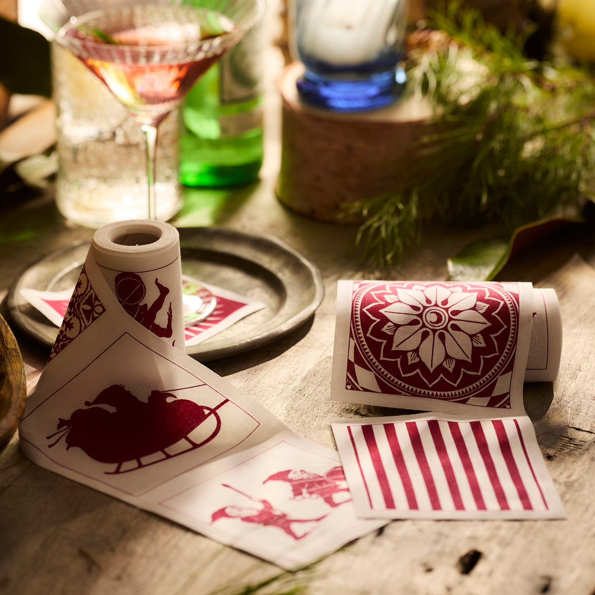 Holiday-themed red and white decorative reusable cocktail cotton napkin rolls are displayed on a table beside a cocktail glass and greenery, accompanied by Caskata&#39;s Holiday Fun Cocktail Napkin Rolls, Set of 2.