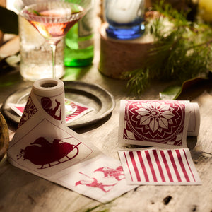 Holiday-themed red and white decorative reusable cocktail cotton napkin rolls are displayed on a table beside a cocktail glass and greenery, accompanied by Caskata's Holiday Fun Cocktail Napkin Rolls, Set of 2.