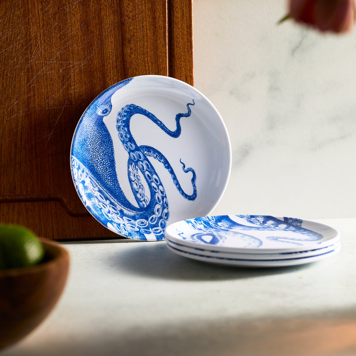 A stack of Caskata&#39;s Lucy Melamine Small Plates, Set of 4, is elegantly placed on a light countertop, complemented by a wooden board and a bowl of limes.