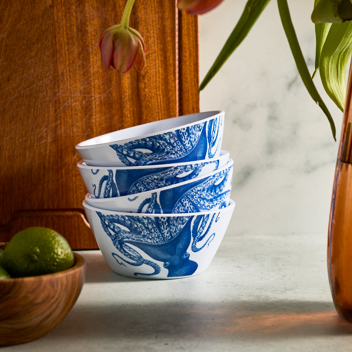 Set of Caskata&#39;s Lucy Melamine Cereal Bowls in white with blue octopus design beside a wooden board, limes in one bowl, and a vase with flowers.