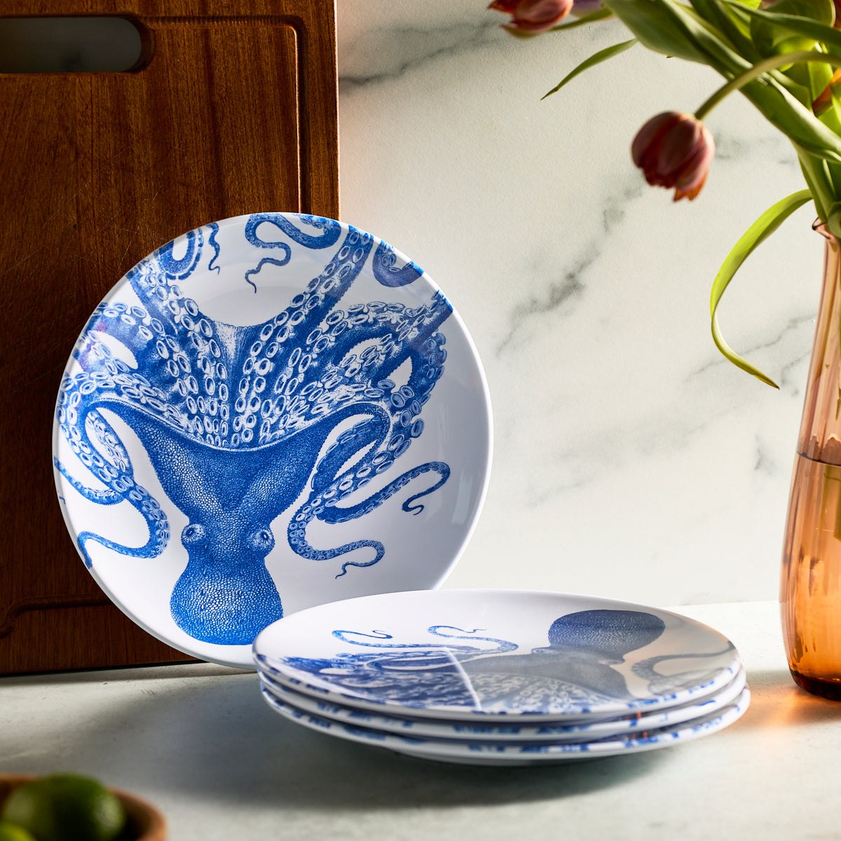 A set of Caskata&#39;s Lucy Melamine Dinner Plates with blue octopus designs is stacked on a countertop next to a wooden board and a vase of tulips.