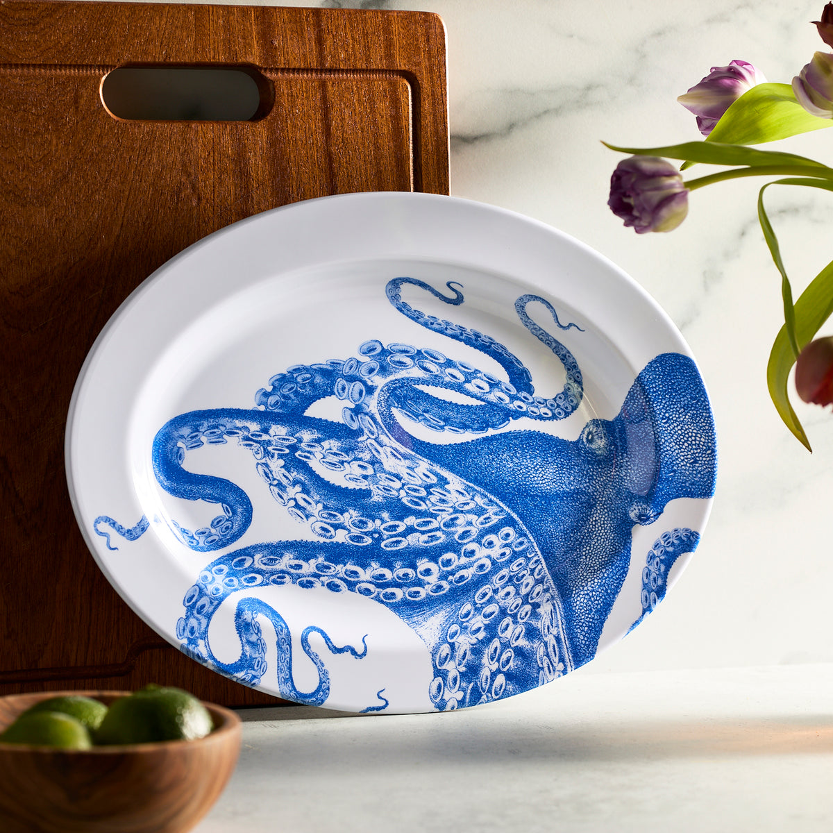 A Lucy Melamine Oval Rimmed Platter by Caskata, set upright against a wooden cutting board, is accompanied by tulips and limes on a marble surface, highlighting the charming unbreakable style in any melamine collection.