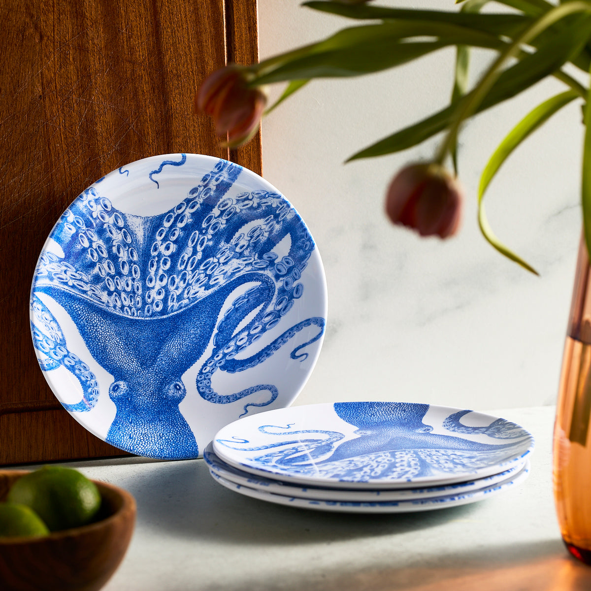 Caskata&#39;s Lucy Melamine Salad Plates, featuring a blue octopus design, stand beside a vase of flowers and a bowl of limes, capturing the whimsical charm of Lucy.