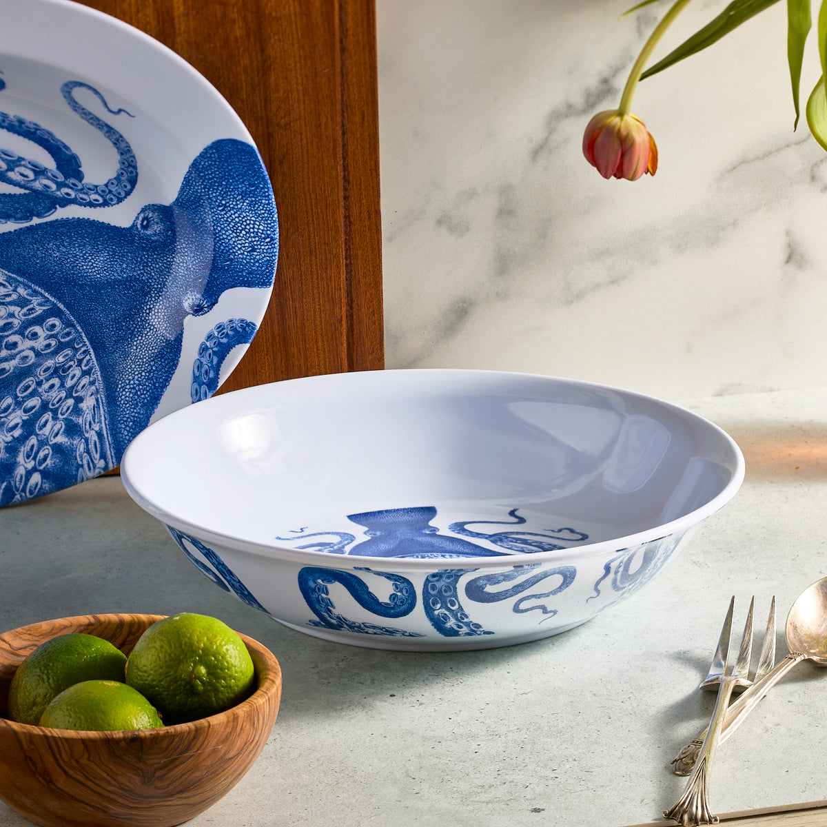 The Lucy Melamine Serving Bowl by Caskata, with its blue octopus design, sits on a marble surface. Nearby, a wooden bowl of limes and an overhead tulip add contrast. A matching plate from the melamine collection completes the scene.