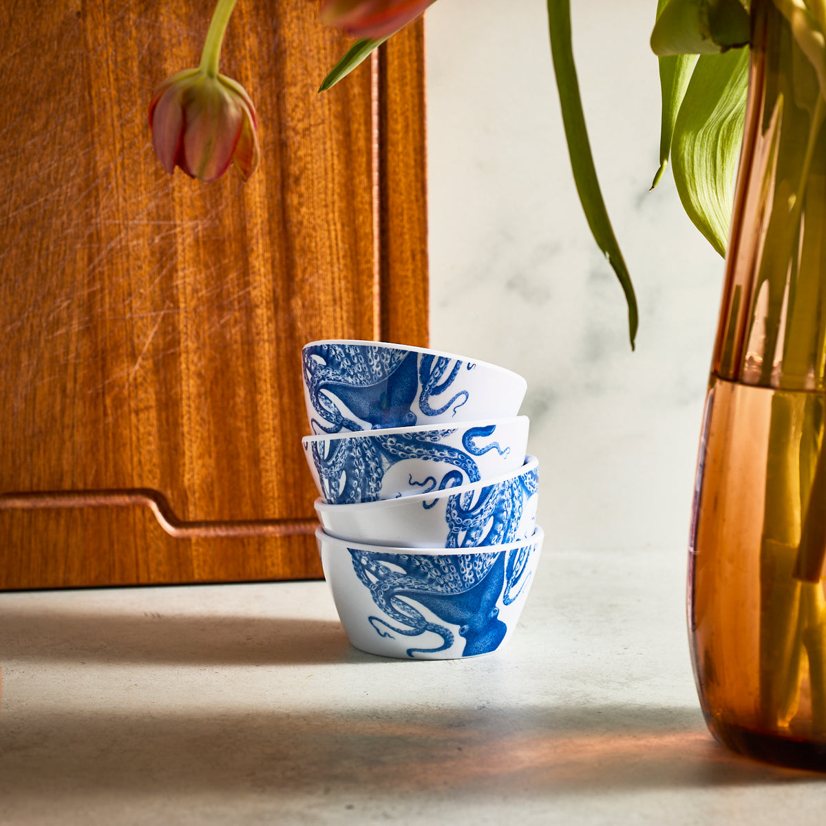 Four stacked Lucy Melamine Snack Bowls by Caskata, featuring a blue and white octopus pattern, are placed beside a copper-colored vase, with a wooden board in the background.