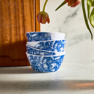 A set of four stacked BPA-free Peony Melamine Cereal Bowls by Caskata, featuring blue floral patterns, are displayed on a marble surface. Nearby, a wooden board adds elegance with pink tulips hanging above, ideal for indoor and outdoor use.