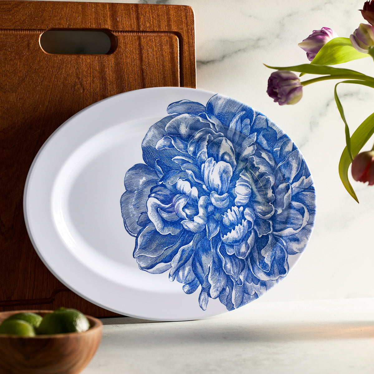 The Caskata Peony Melamine Oval Rimmed Platter, featuring a large blue flower design, is displayed vertically against a wooden cutting board. Delicate tulips and fresh limes complete the elegant setting of this unbreakable dinnerware, perfect for any occasion.