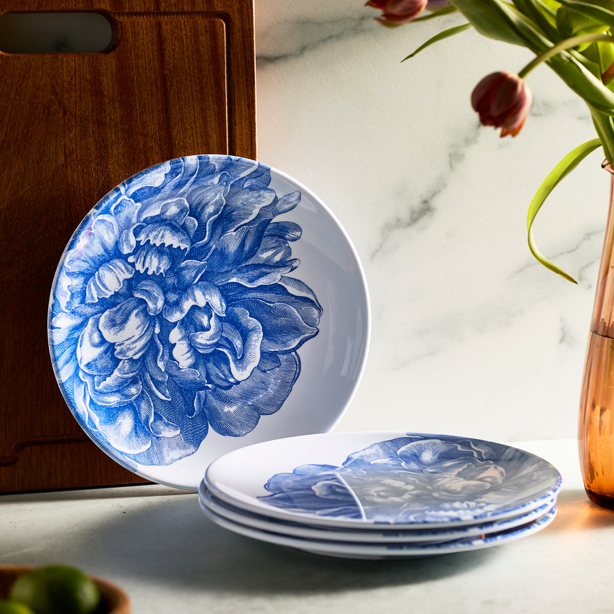 A stack of Caskata&#39;s Peony Melamine Dinner Plates, Set of 4, featuring a blue floral design, stands upright against wood and marble. Tulips in a vase enhance the scene’s charm, evoking an unbreakable style.