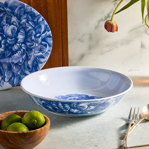 The Peony Melamine Serving Bowl by Caskata, featuring a blue floral pattern, rests on the countertop. Nearby are a wooden bowl of limes, a fork, and a knife. A tulip leans into the scene while in the background, an elegant matching plate completes this unbreakable style tableau.