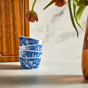 On a stone surface, stacked blue and white floral-patterned bowls feature near a wooden board and tulips. These elegant Peony Melamine Snack Bowls by Caskata combine unbreakable style with durability, crafted from BPA-free melamine dinnerware in a set of 4.