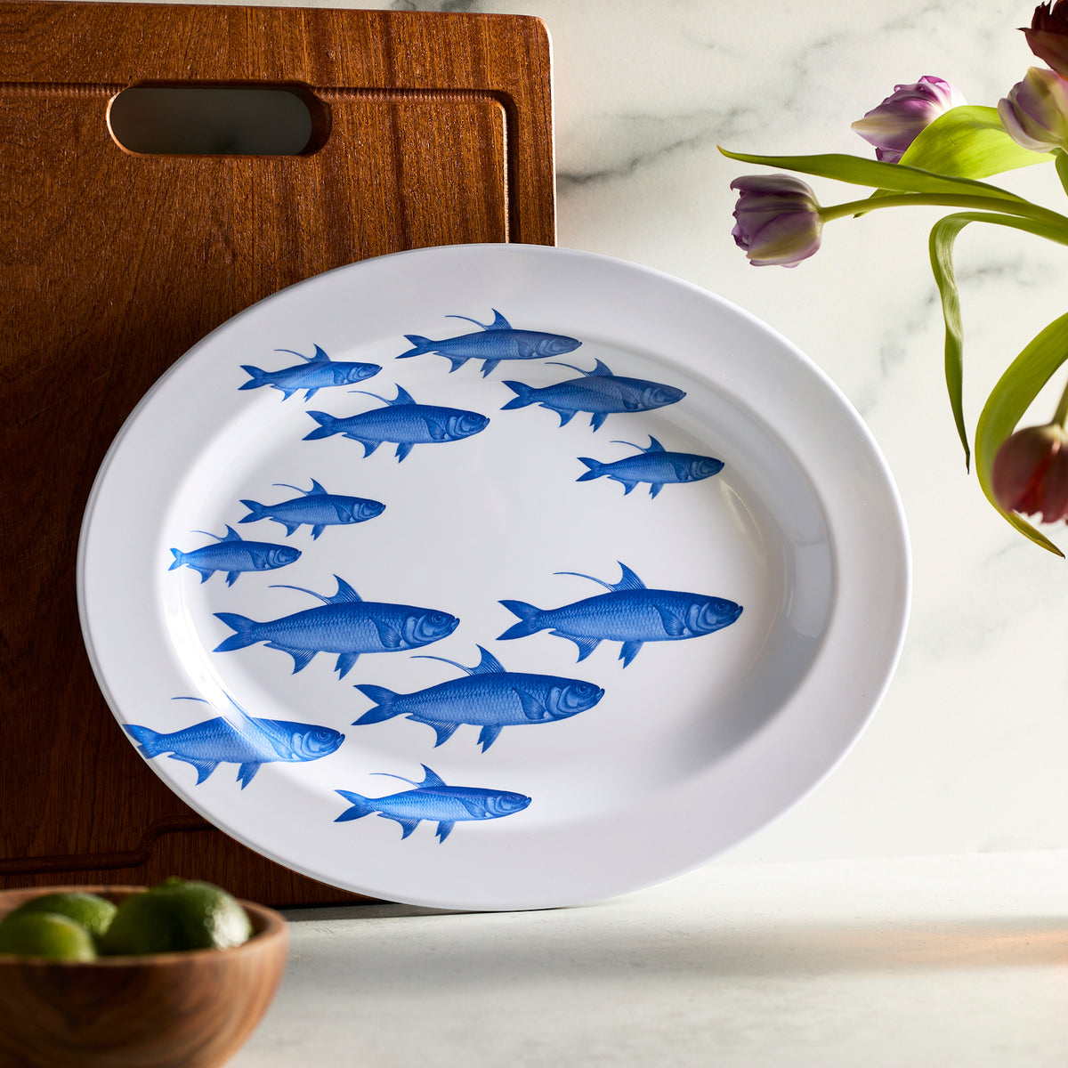 The Caskata School of Fish Melamine Oval Rimmed Platter, part of our outdoor collection, leans against a wooden cutting board. BPA-free and ideal for alfresco dining, it&#39;s paired with vibrant tulips and limes to add charm.