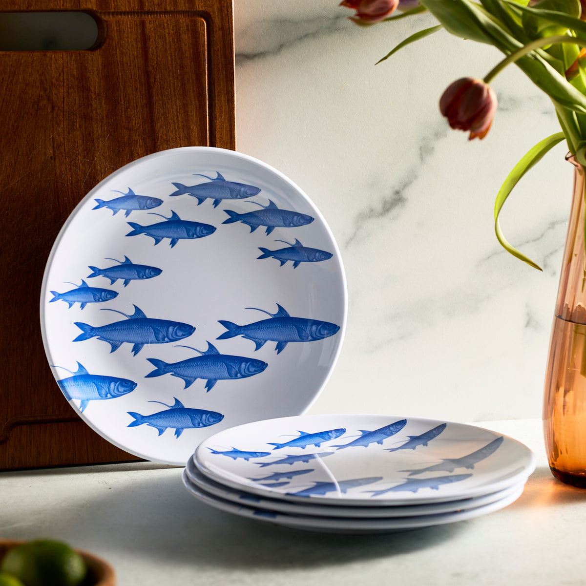 A set of Caskata’s School of Fish Melamine Dinner Plates, featuring blue fish designs, sits on a marble surface beside a wooden cutting board and a tulip-filled vase.