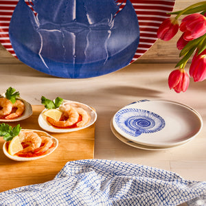 Three Caskata Artisanal Home Shells Small Plates with shrimp, lemon, and parsley sit on a wooden board beside stacked plates. A blue-patterned napkin and red tulips are nearby. In the background is an heirloom-quality plate featuring a delicate lobster design from our Shells collection.