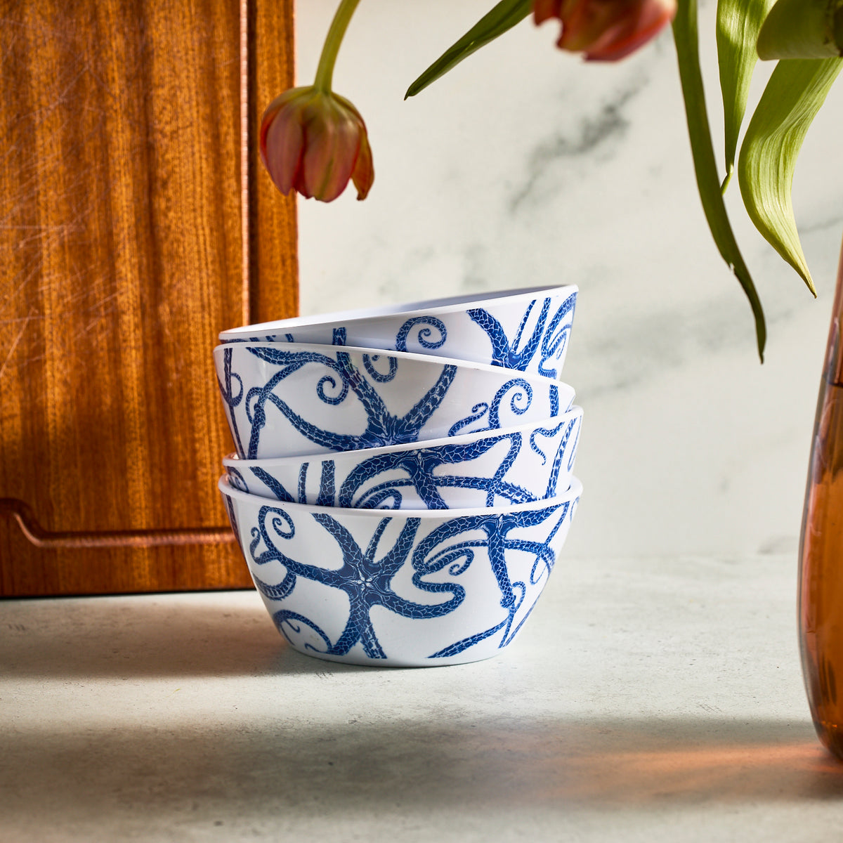 A set of four Caskata Starfish Melamine Cereal Bowls, featuring BPA-free white with blue abstract starfish patterns, sits on a countertop beside a wooden board and a vase with drooping red flowers.